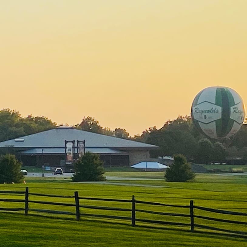 A large decorative hot air balloon near a building at sunset, with green grass and a fence in the foreground.