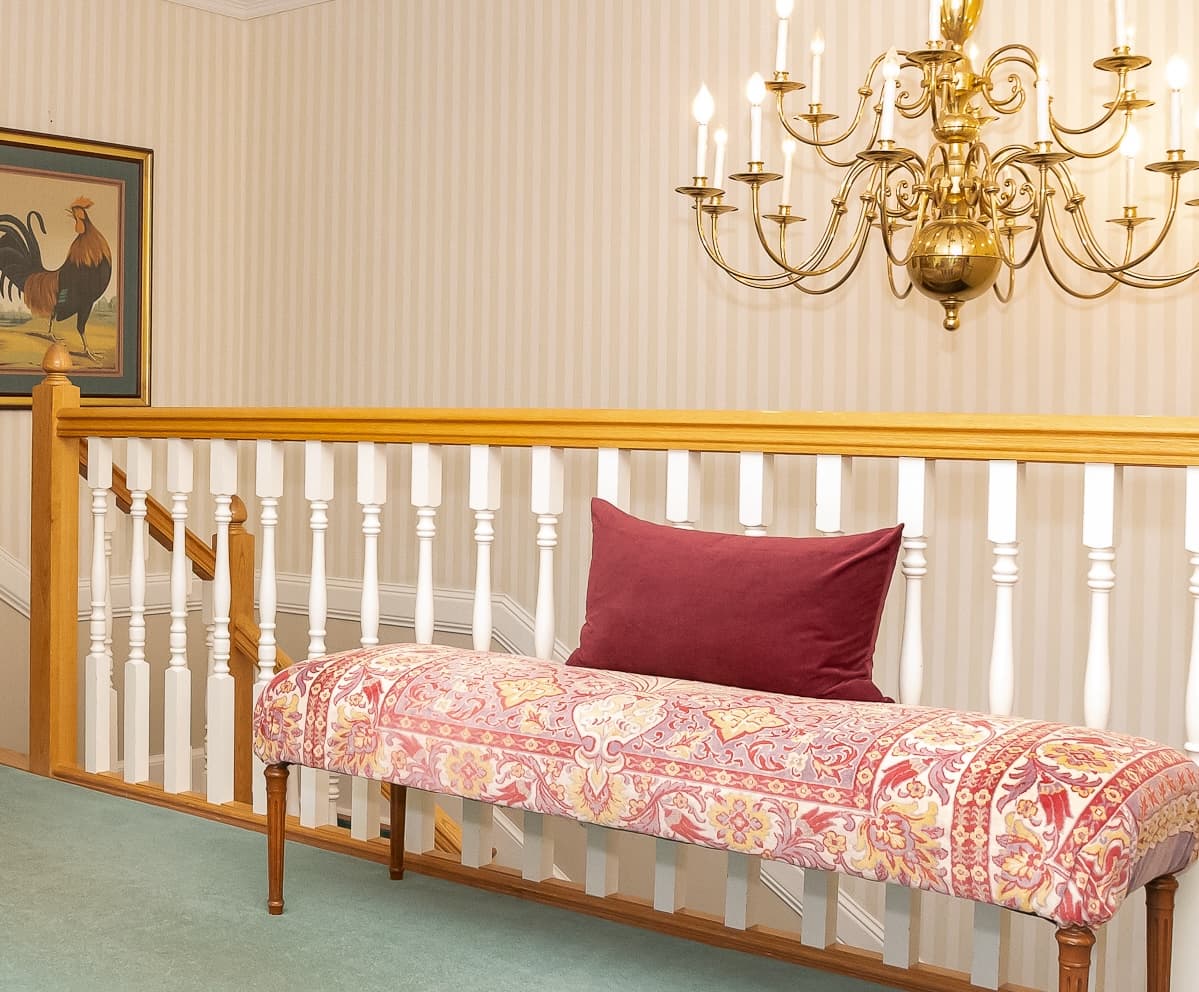 A patterned bench with a burgundy pillow is situated near a chandelier and a staircase in a softly lit hallway.