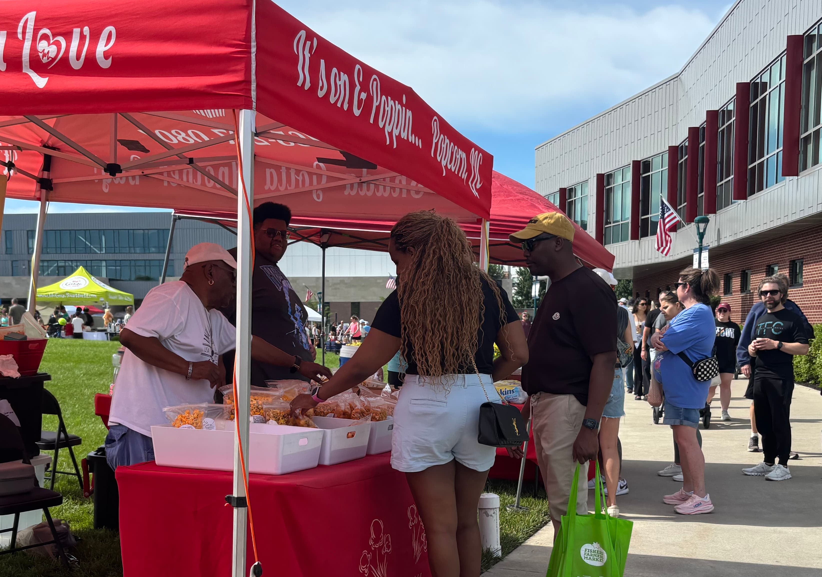 A diverse group of people gathers at a vendor stand under a red tent, sampling popcorn at an outdoor event.