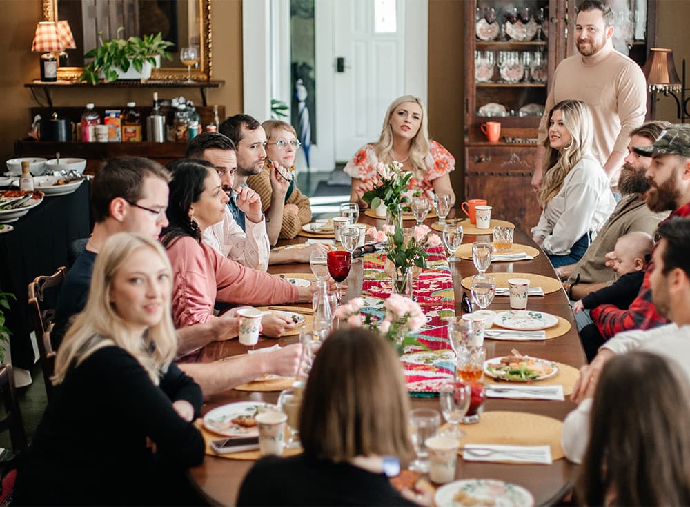 A large group of people sits around a long dining table filled with food and drinks, engaged in conversation.