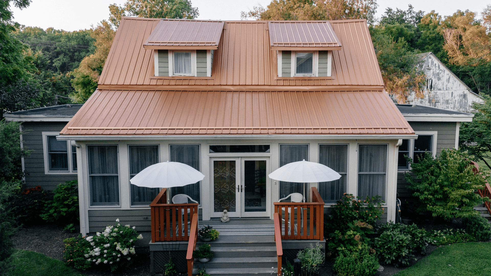 A charming house with a copper roof, featuring a front porch and umbrellas on either side, surrounded by greenery.