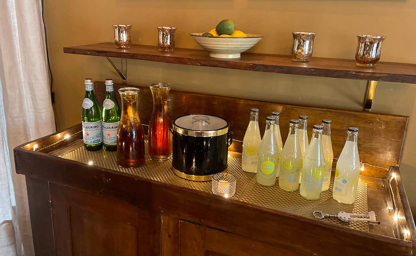 A wooden bar cart displays various drinks, including sparkling water, iced tea, and lemonade, along with a bowl of fruit on the shelf above.