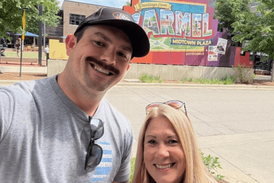 A man and a woman smile for a selfie in front of a colorful mural that says "Greetings from Carmel" at Midtown Plaza.