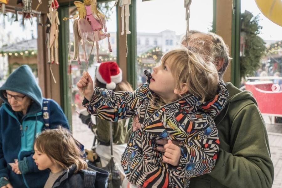 A child points at hanging decorations while being held by an adult, with other people in the background.