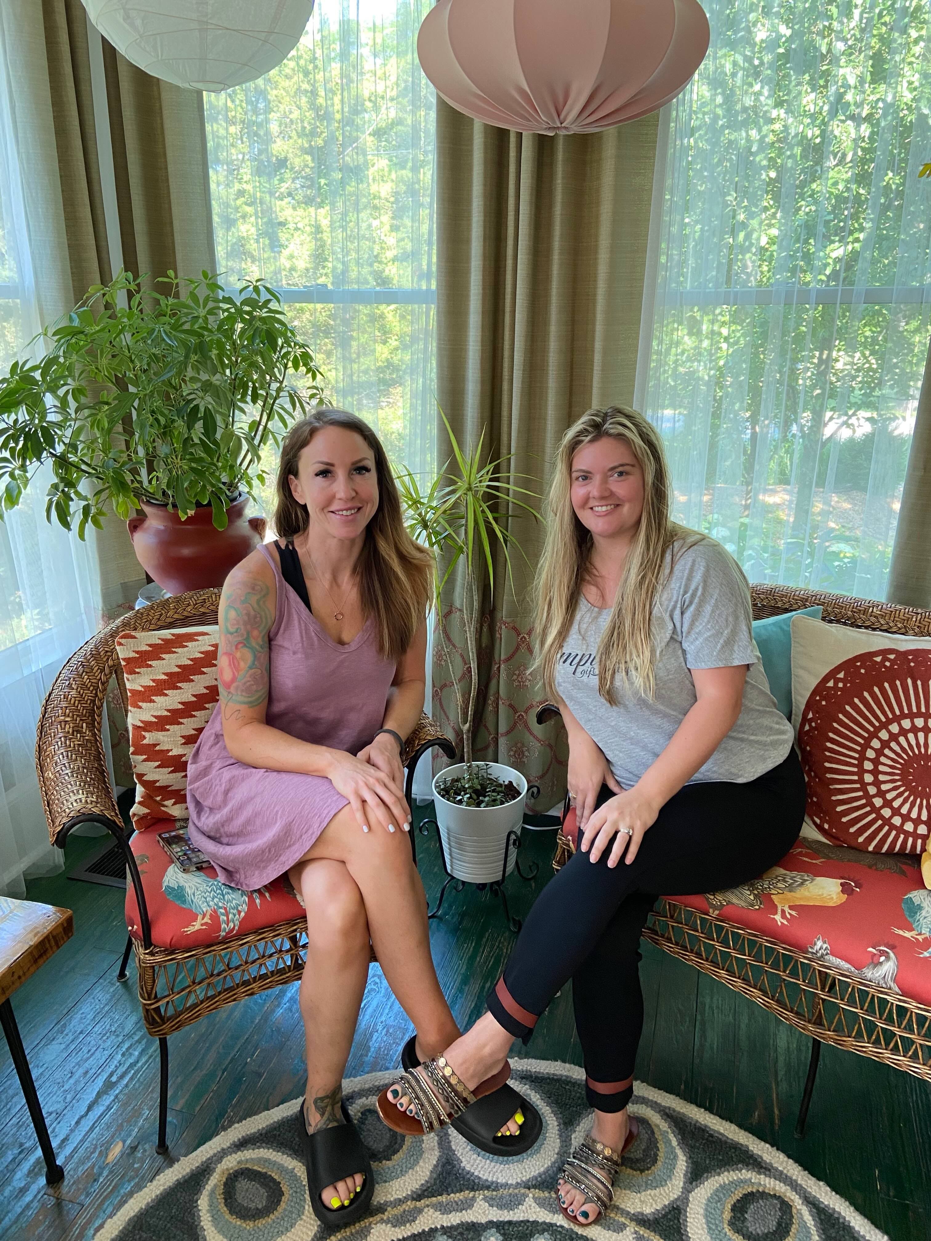 Two women sit together on a porch, surrounded by plants and colorful decor.