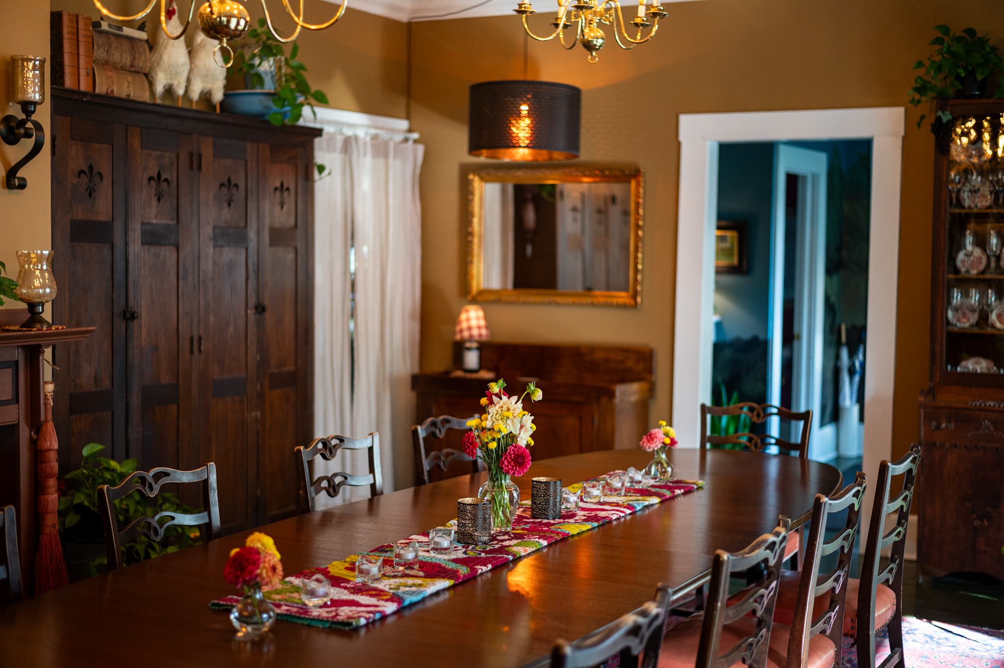 A beautifully set dining room with a long wooden table adorned with flowers and a colorful runner, surrounded by antique furniture.