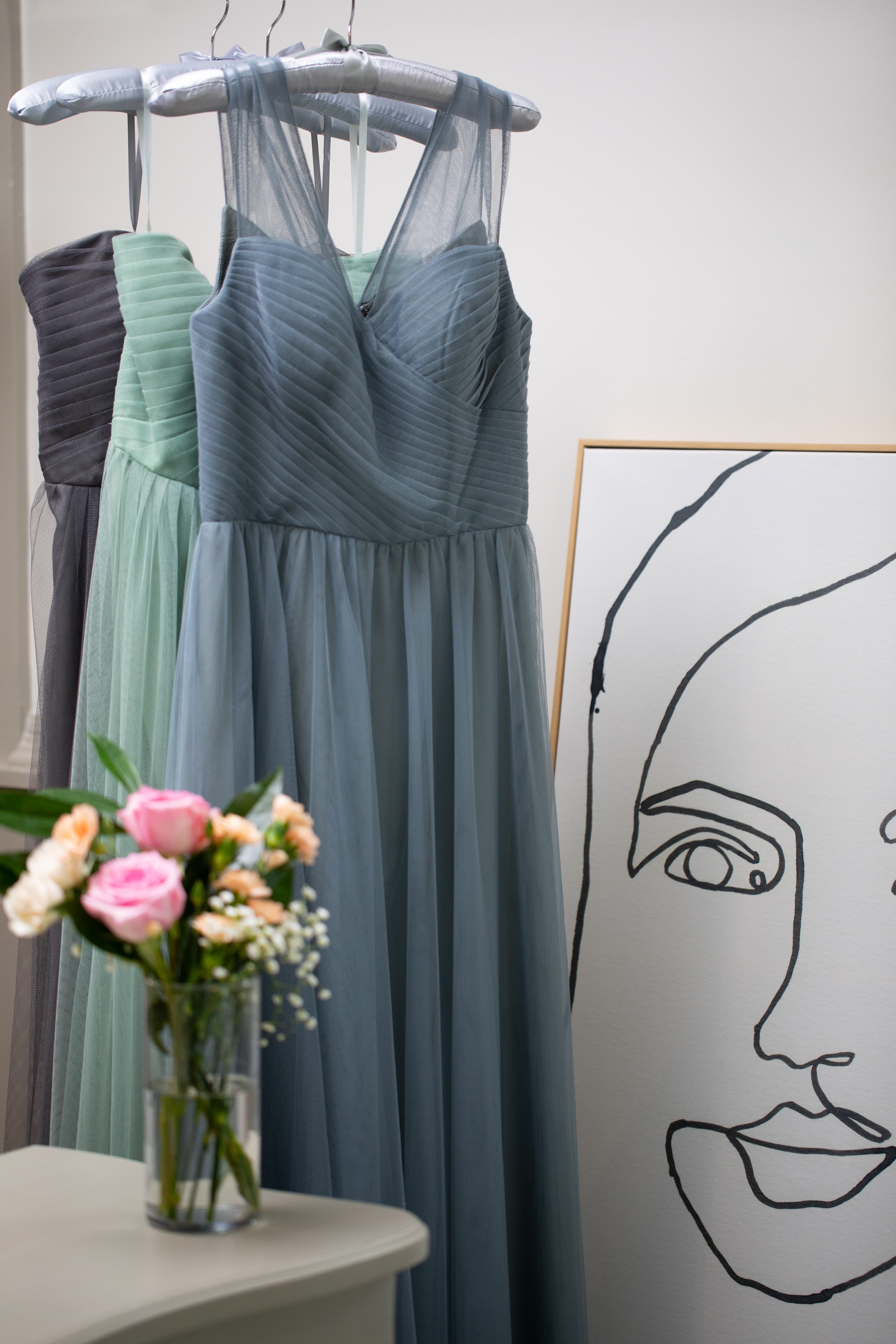 Three elegant dresses in soft colors hang beside a minimalist line drawing of a face and a vase of flowers.