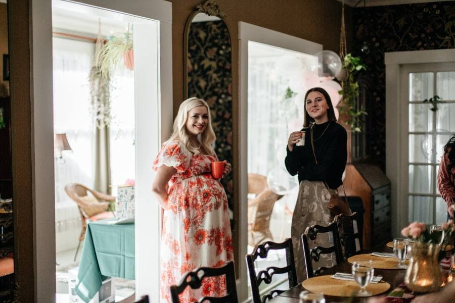 Two women stand in a warmly decorated room, one holding a cup and the other a small red mug, while seated tables and greenery are visible in the background.