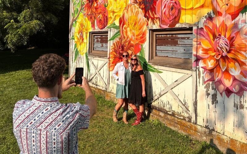 Two women pose for a photo in front of a colorful flower mural while a man takes their picture.