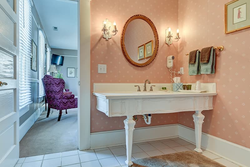 A vintage bathroom featuring a white vanity with a circular mirror and pink patterned walls, visible through an opened doorway to a cozy sitting area.