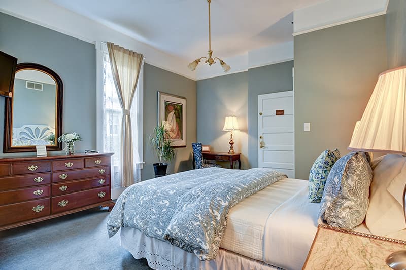 A cozy bedroom featuring a bed with decorative pillows, a wooden dresser, a painting on the wall, and large windows with curtains.