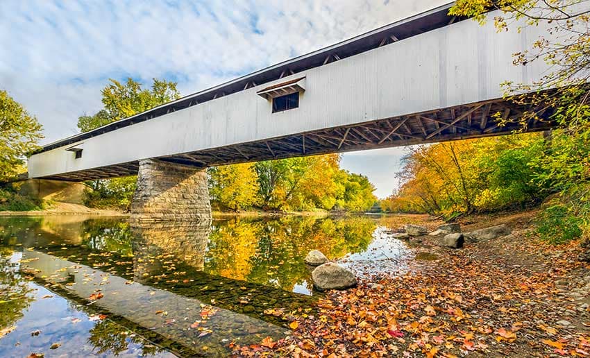 A white covered bridge spans a calm river framed by vibrant autumn foliage. A white covered bridge spans a calm river framed by vibrant autumn foliage.