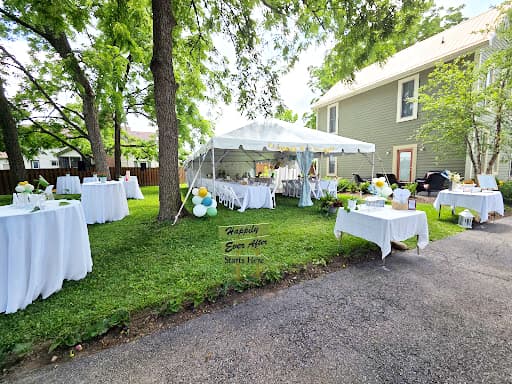 A festive outdoor setup with a white tent and decorated tables, surrounded by greenery and a house.