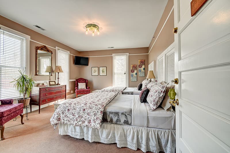 A cozy bedroom featuring two beds with floral bedding, a dresser, a mirror, and ample natural light from nearby windows.