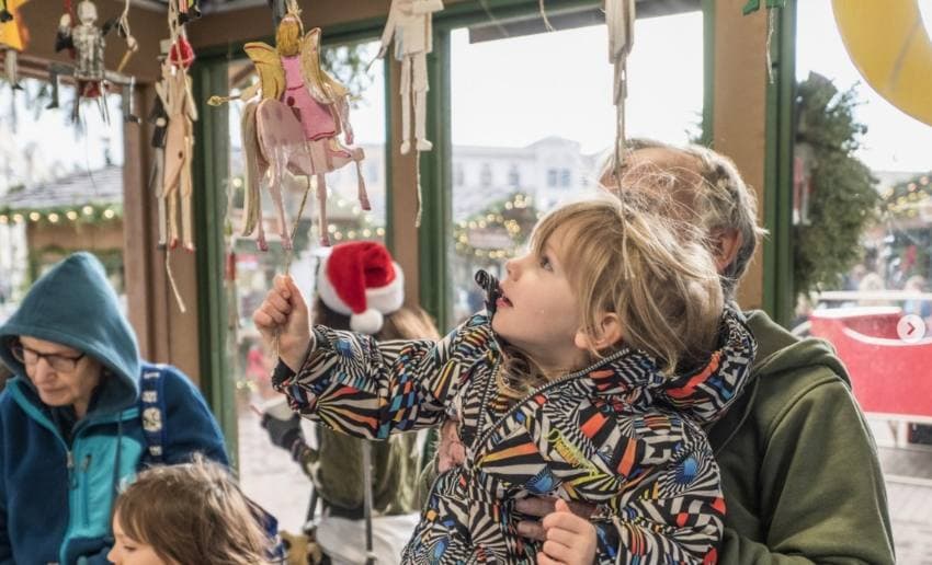 A young child curiously reaches for a hanging ornament while adults watch nearby in a festive setting. A young child curiously reaches for a hanging ornament while adults watch nearby in a festive setting.