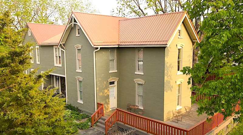 A two-story house with a green exterior and a red metal roof, surrounded by trees.