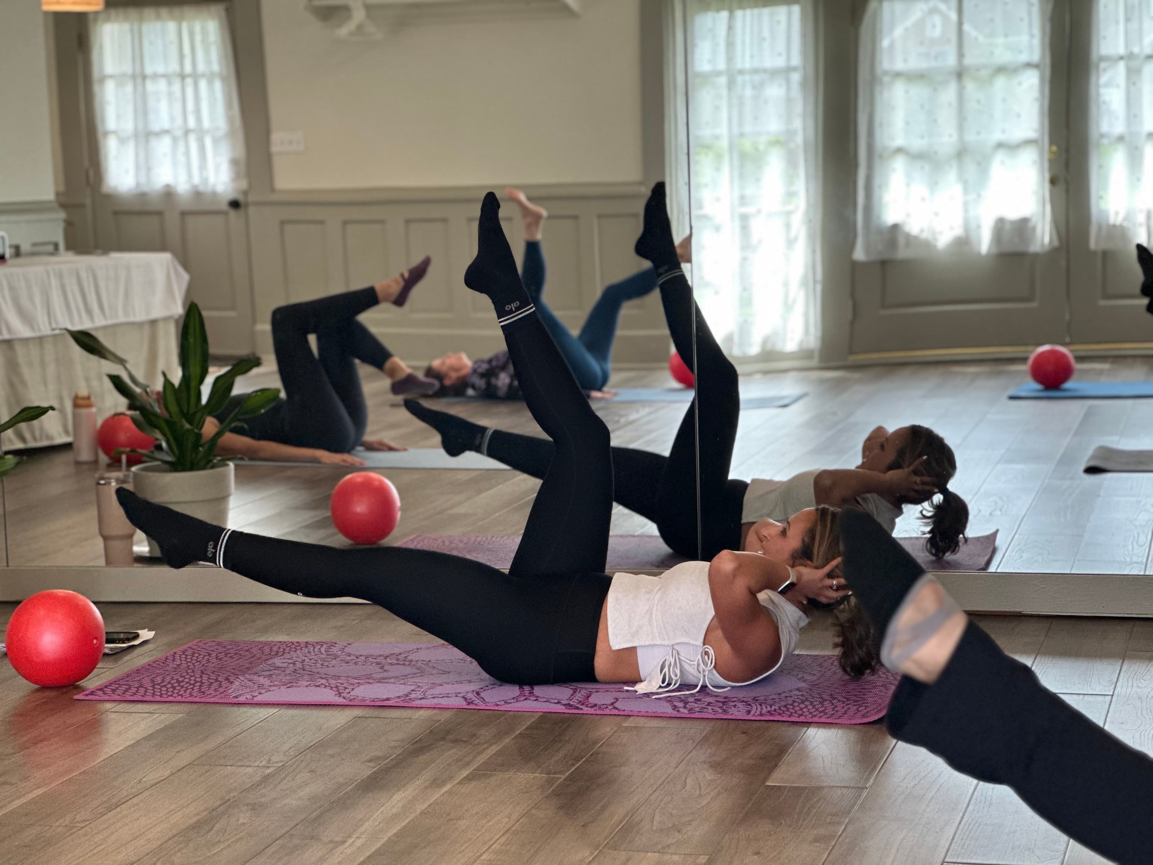 Two women in leggings do Pilates exercises on mats in a mirrored studio. Two women in leggings do Pilates exercises on mats in a mirrored studio.