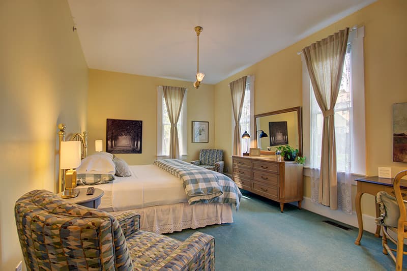 A cozy bedroom featuring a bed with a checkered blanket, two armchairs, and large windows with light curtains.