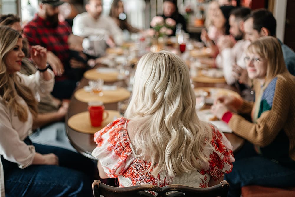 A group of people enjoying a meal around a table, with focus on a woman with long, wavy hair facing away from the camera.