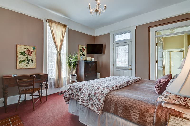 A cozy bedroom featuring a floral-patterned bedspread, a wooden desk, and large windows with sheer curtains.