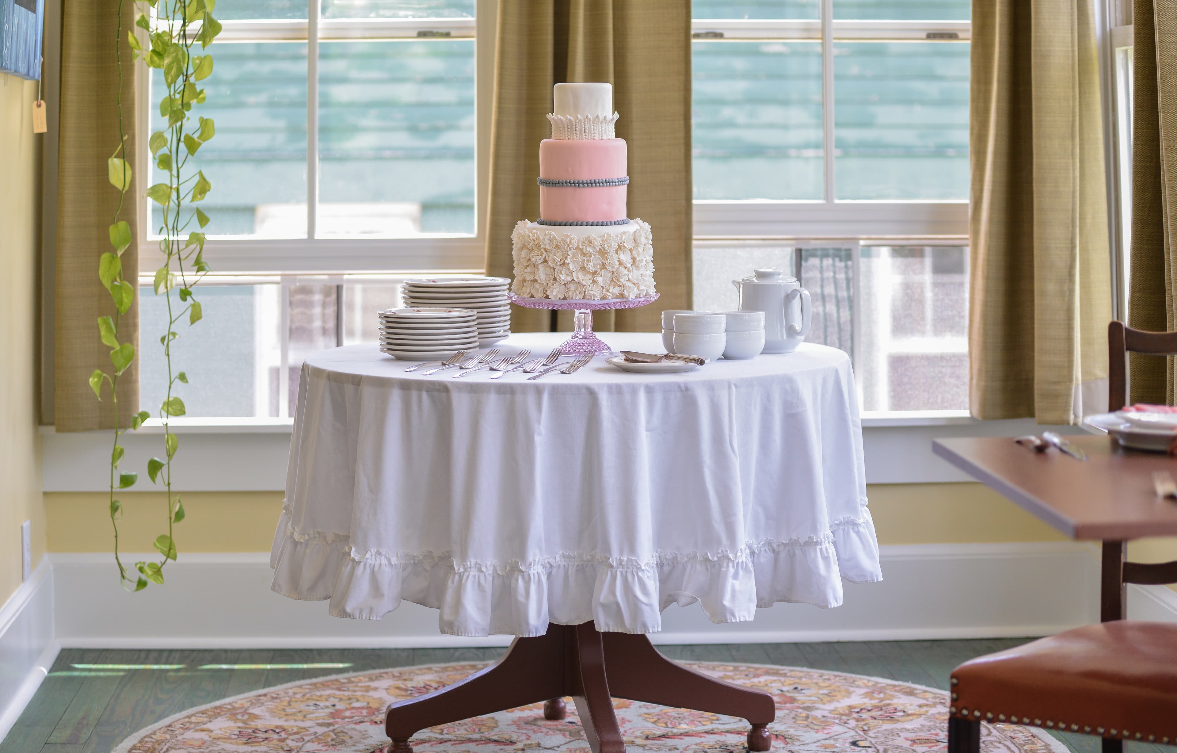 A round table dressed in a white tablecloth with a tiered pink cake, plates, and cups, set beside a window with green curtains.