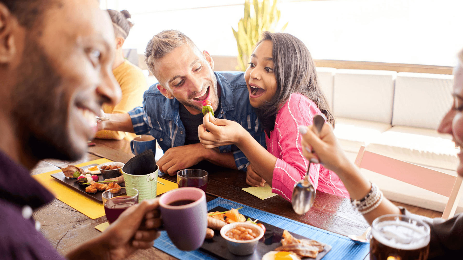 A group of four people joyfully sharing a meal at a table, with one person feeding another.