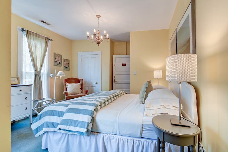 A cozy bedroom with yellow walls, a bed featuring striped linens, and a chandelier.