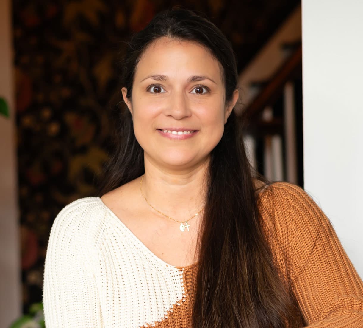 Smiling woman with long dark hair wearing a two-toned sweater, standing indoors.