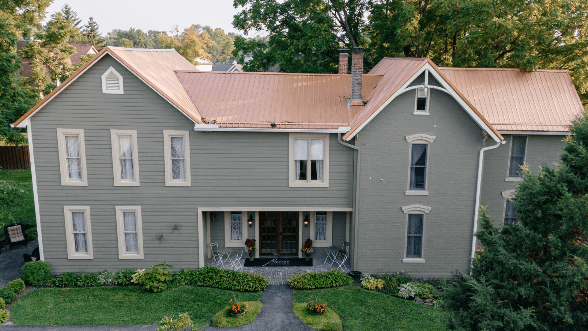 A large, two-story house with a copper roof, surrounded by greenery and flower beds.