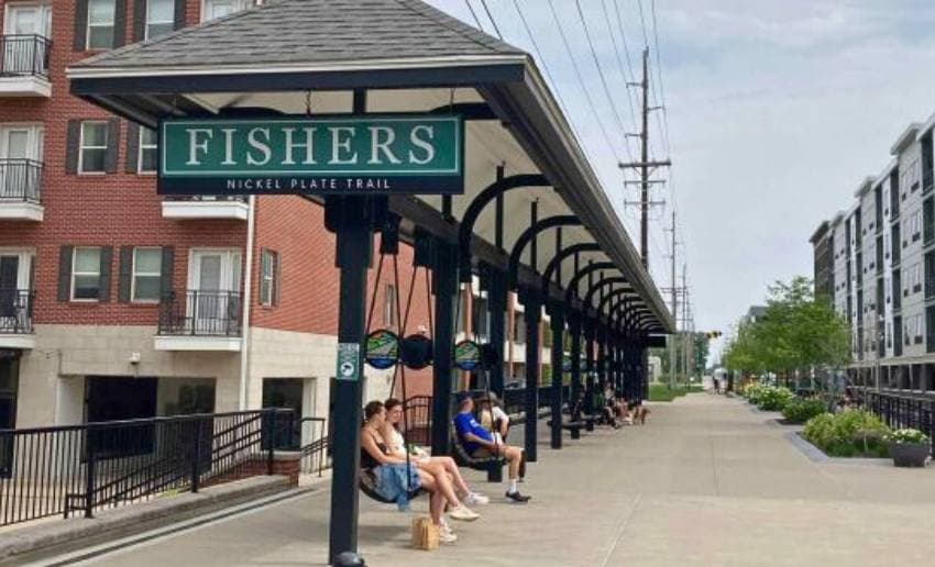 A sign for the Fishers Nickel Plate Trail stands next to a seating area where people relax. A sign for the Fishers Nickel Plate Trail stands next to a seating area where people relax.