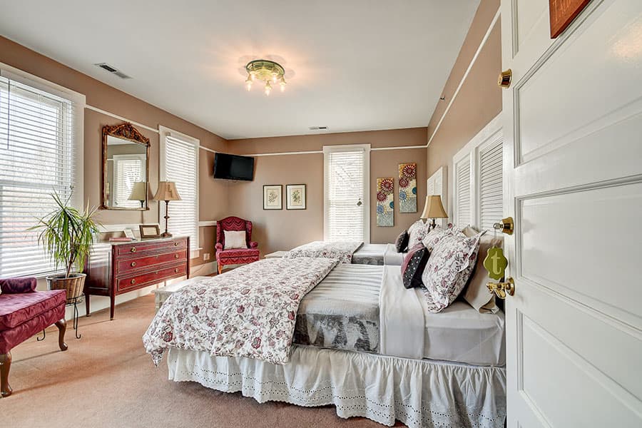 A cozy bedroom featuring twin beds, a vintage dresser, large windows with natural light, and decorative accents.