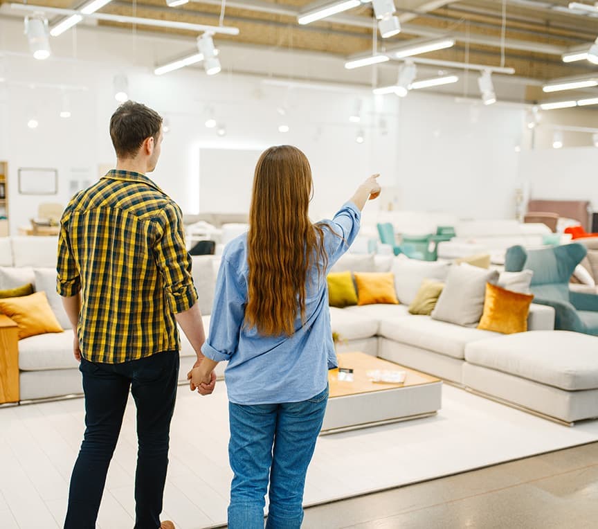 A couple looks at furniture in a showroom, with the woman pointing towards a sofa.
