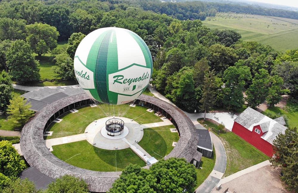 A colorful hot air balloon with "Reynolds" branding floats above a circular park surrounded by trees and a barn.