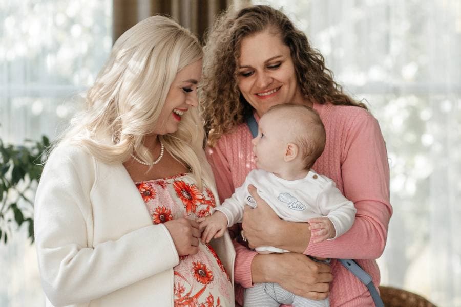 A pregnant woman and a mother smile at each other while holding a baby in a bright, cozy setting.