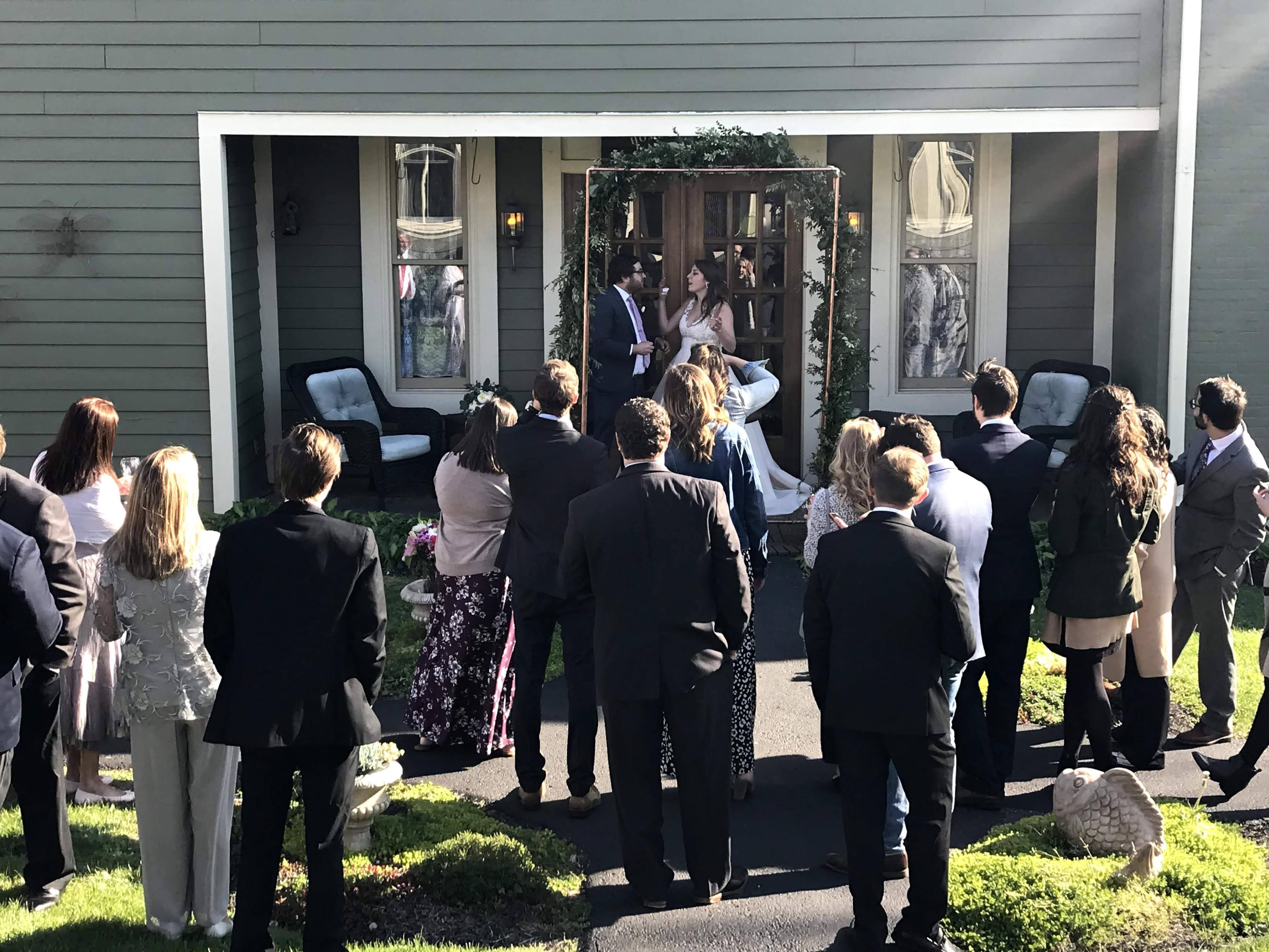 A wedding couple stands at the entrance of a house, surrounded by guests watching the ceremony.
