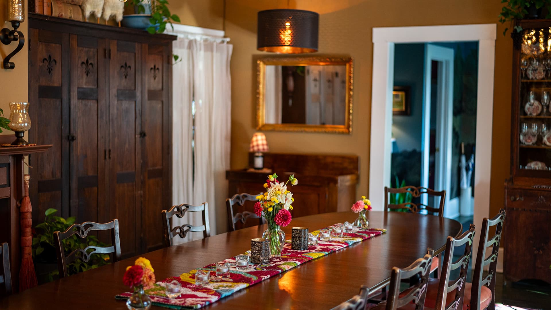 A warmly lit dining room featuring a long wooden table adorned with flowers and a colorful runner, surrounded by chairs and vintage decor.
