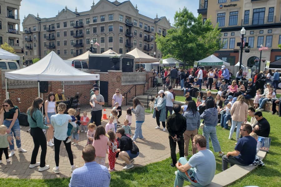 A busy outdoor market event with families and vendors under white tents in a park-like setting.