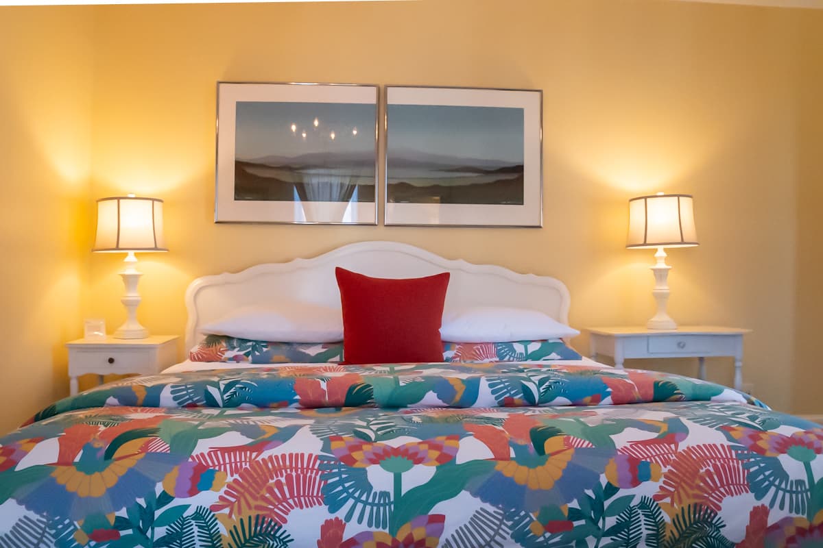 A cozy bedroom featuring a colorful floral comforter, two bedside lamps, and framed artwork on a yellow wall.