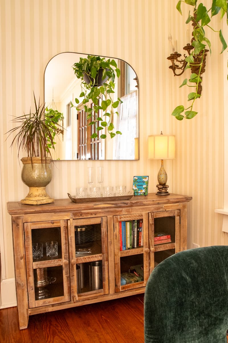 A wooden sideboard displays glasses and books under a round mirror, with plants and a lamp above.