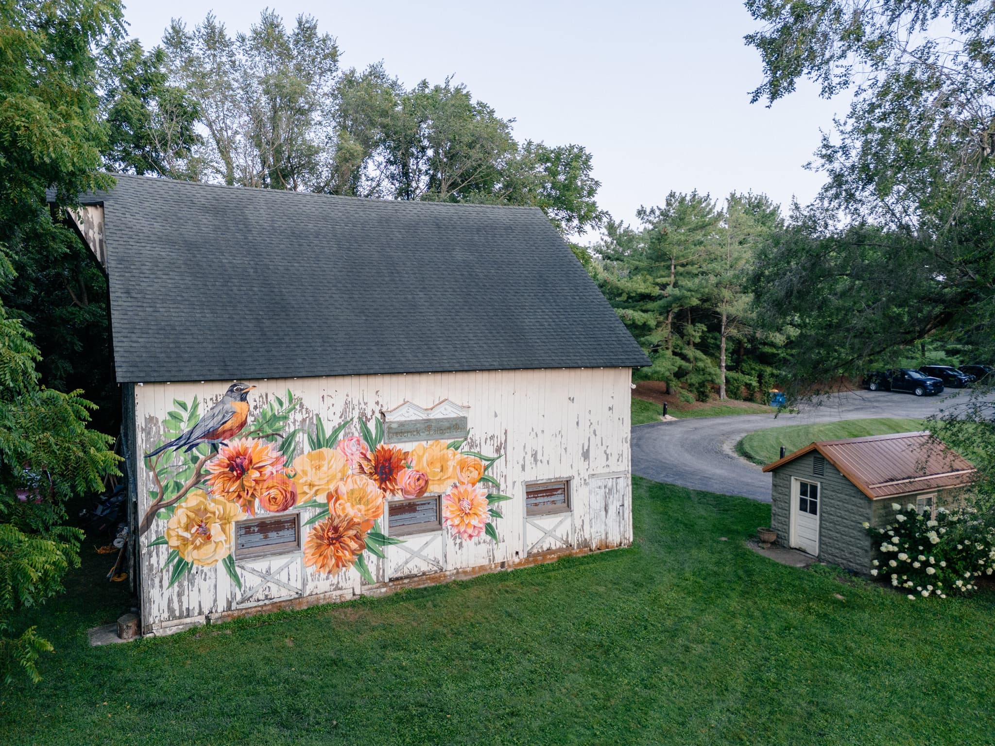 A colorful mural of flowers decorates a white barn surrounded by trees and a nearby shed.