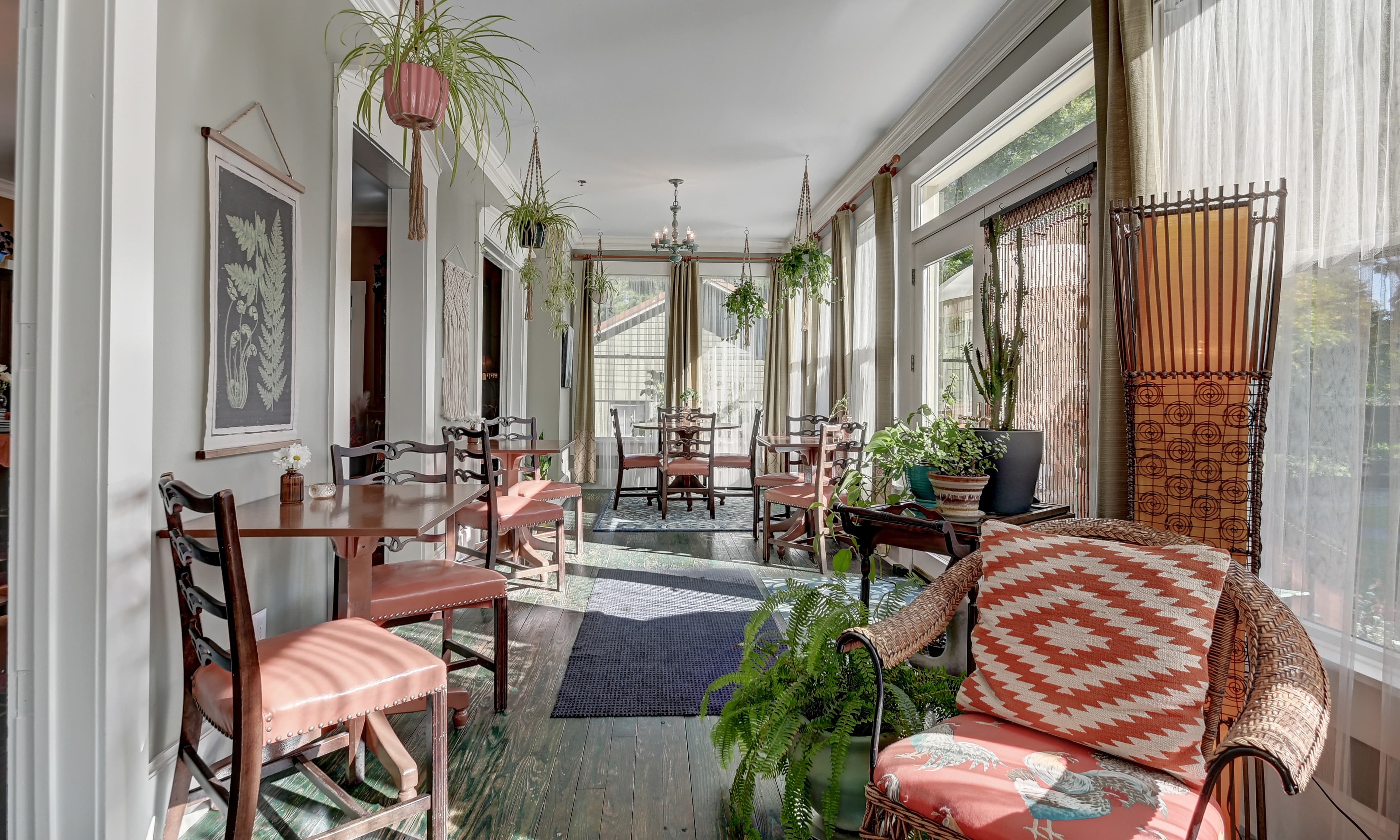 A bright, airy sunroom with plants, wooden tables, and cozy seating.