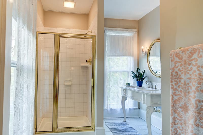 A bright, modern bathroom featuring a glass shower, vanity with a round mirror, and a potted plant by the window.