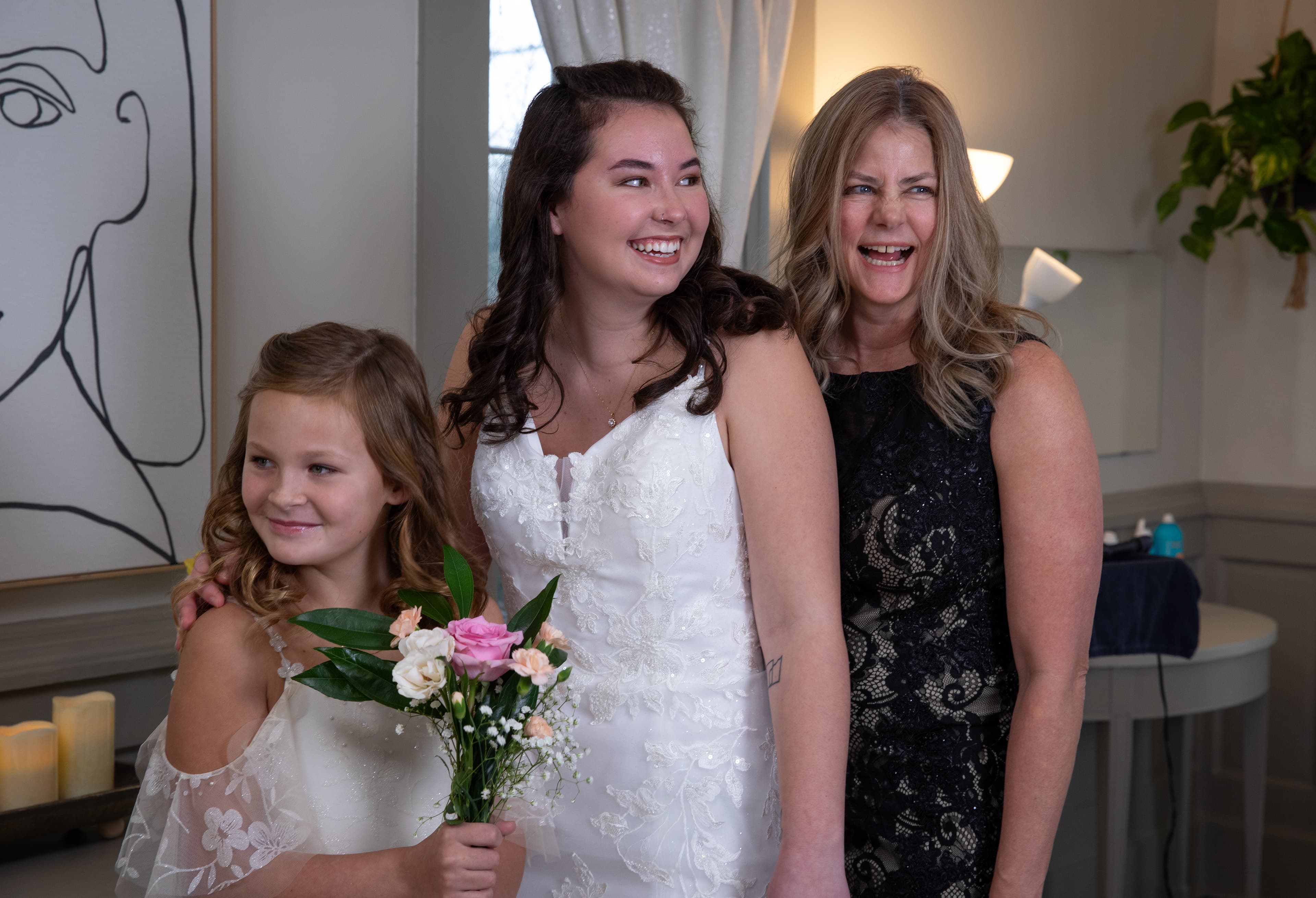 A bride stands happily with her mother and a young girl, all smiling in a warmly lit room.