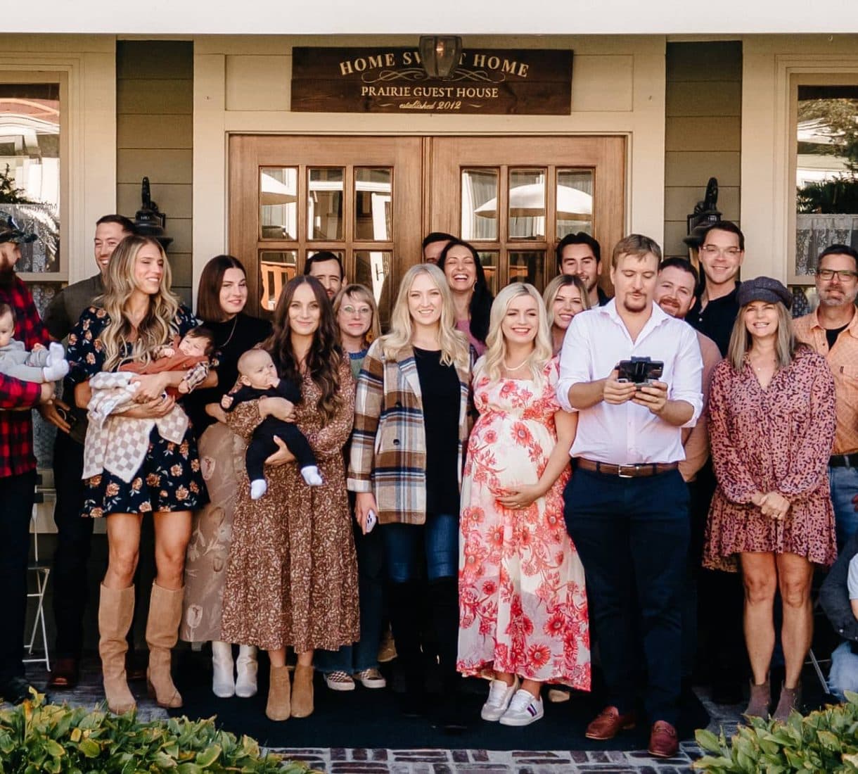 A large group of people, including several holding babies, stands together in front of a guest house, smiling for the camera.