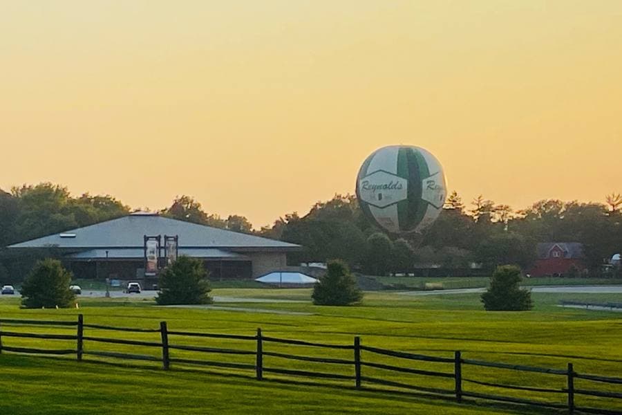 A large green and white soccer ball-shaped hot air balloon hovers above a park with a building and trees at sunset.