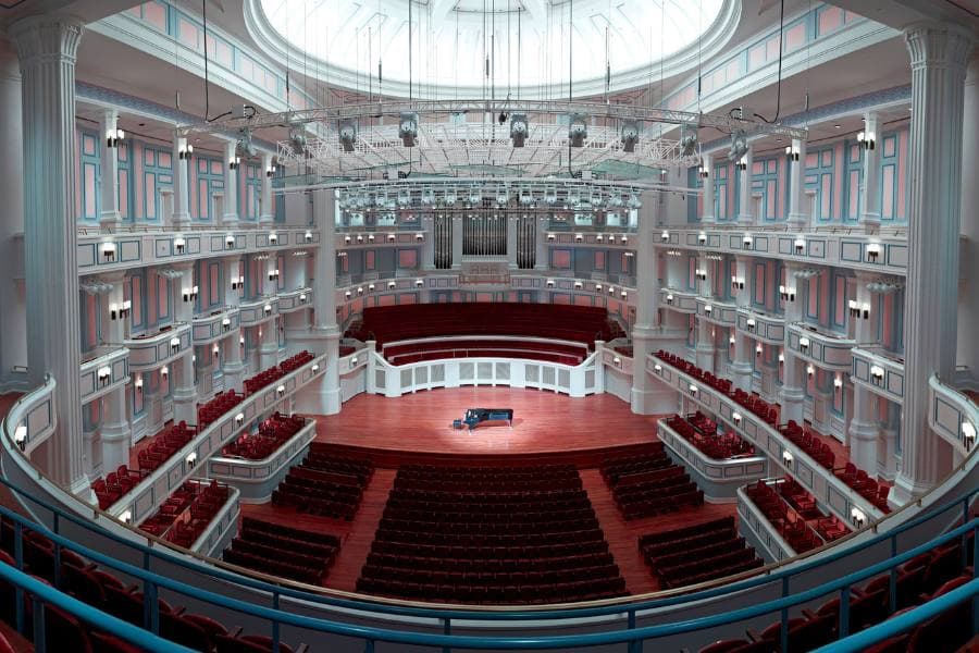 A grand concert hall featuring tiered seating, ornate columns, and a piano on stage.
