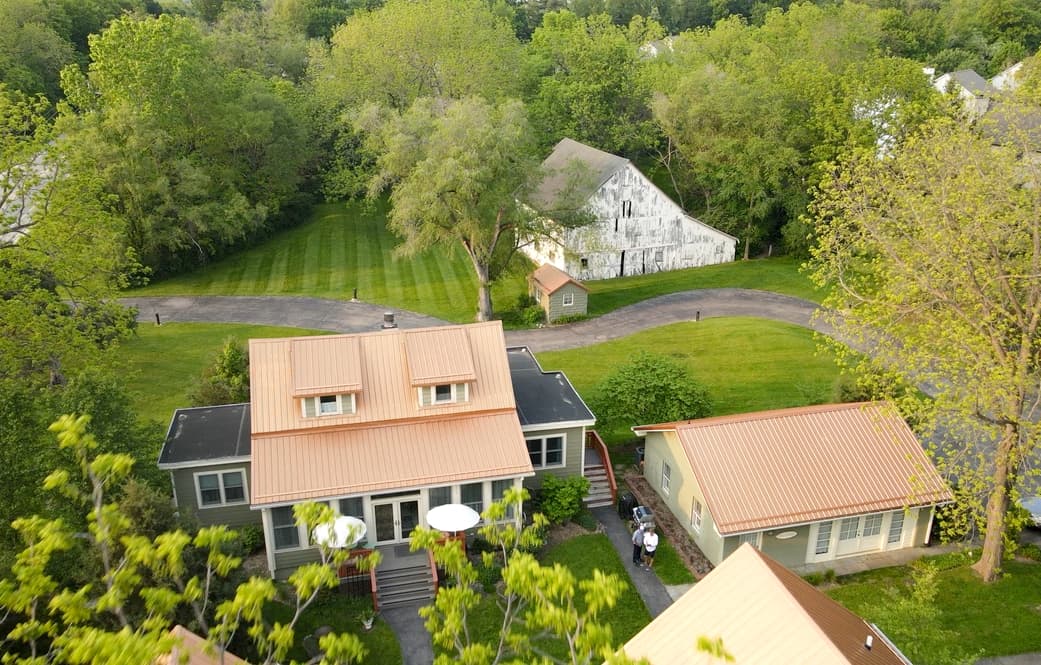 Aerial view of a green landscape featuring a modern house with a metal roof and an old barn in the background.