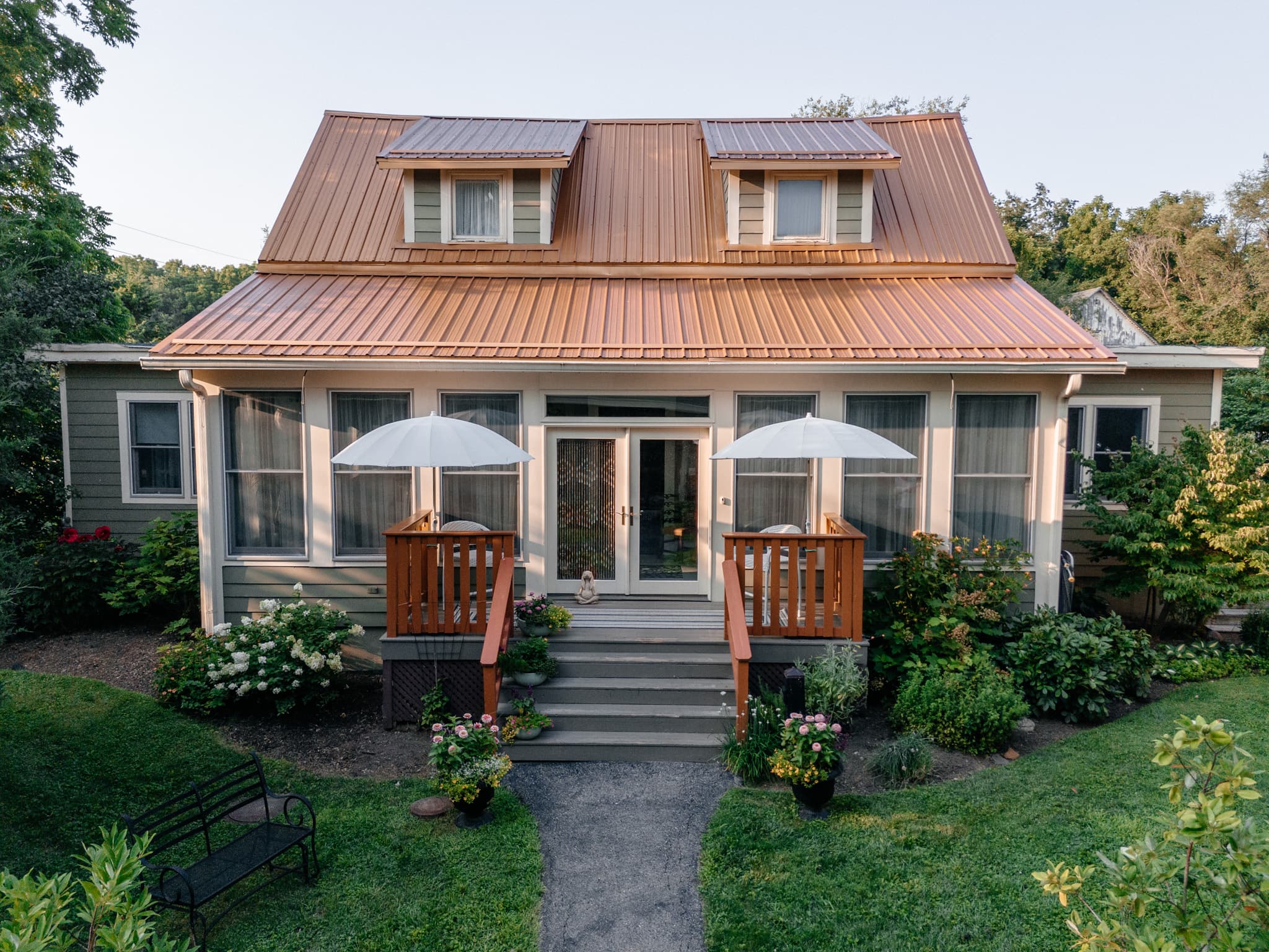 A picturesque house with a copper roof, featuring two front porches with umbrellas and surrounded by lush greenery.