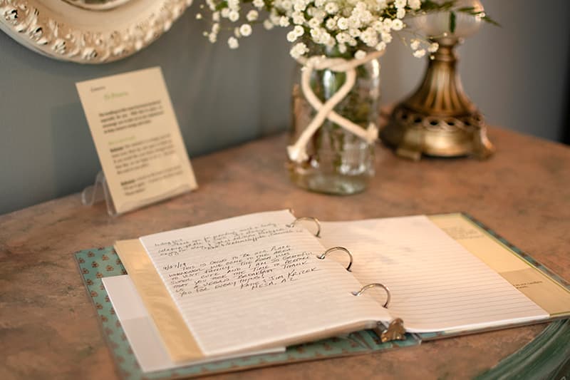 An open notebook with handwritten notes sits on a table next to a vase of flowers and a small sign.