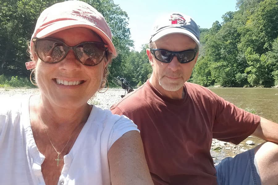 A smiling woman and man sit by a river with trees in the background and a dog nearby.